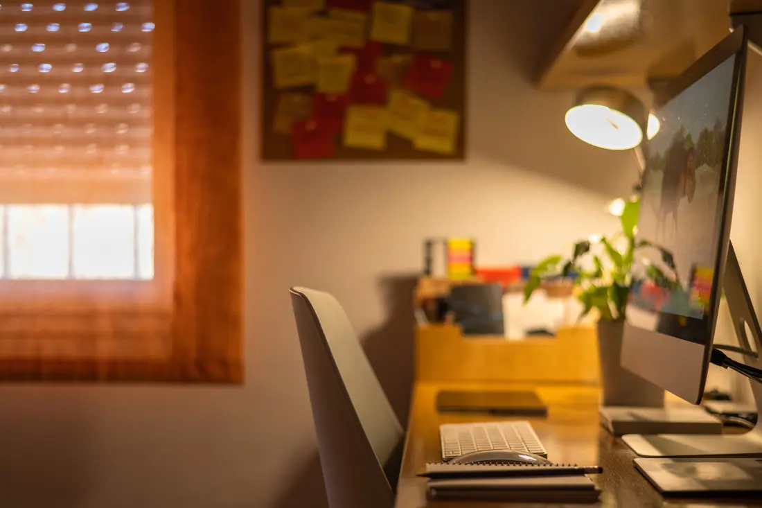A desk and monitor in a home office with the blind drawn against the sun