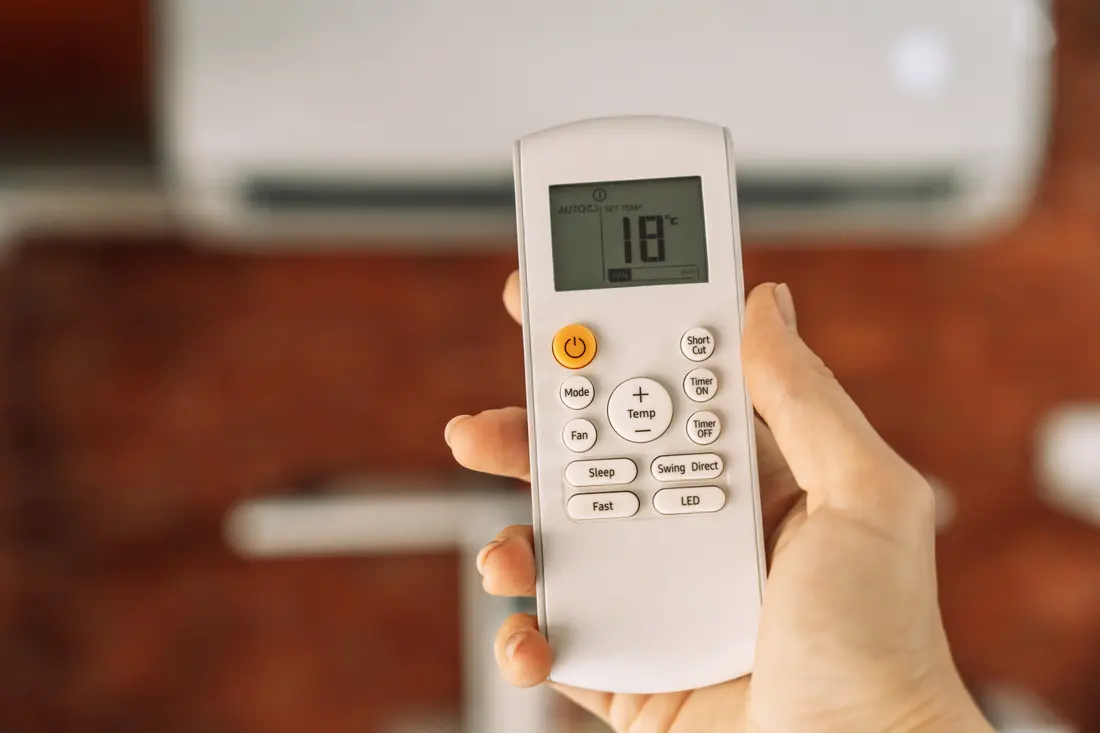 A hand using an air conditioning remote control in front of a wall-mounted unit
